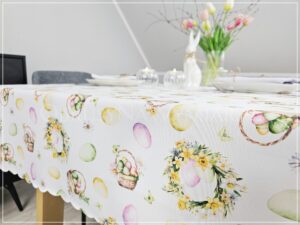 Dekoracja stołu wielkanocnego: biały ceramiczny zając i świece na obrusie z motywem wiosennych wianków.